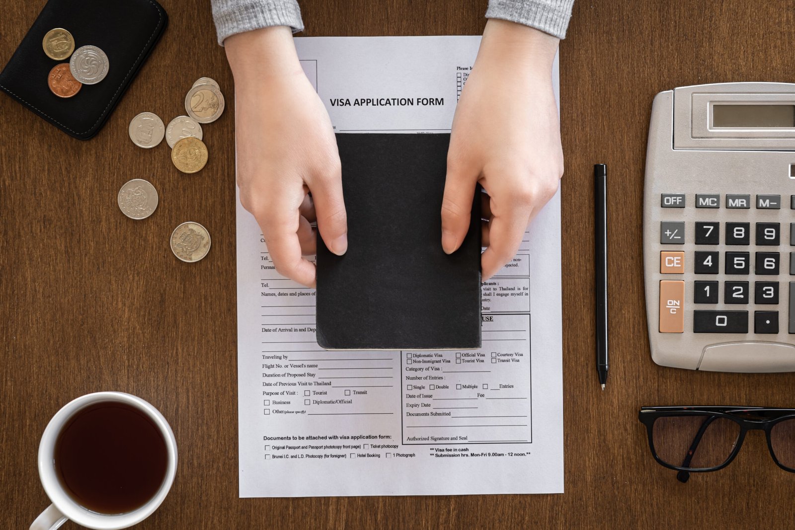 Visa application form, passport in female hands, coins, calculator, coffee on a wooden background, flat lay, visa processing, immigration.